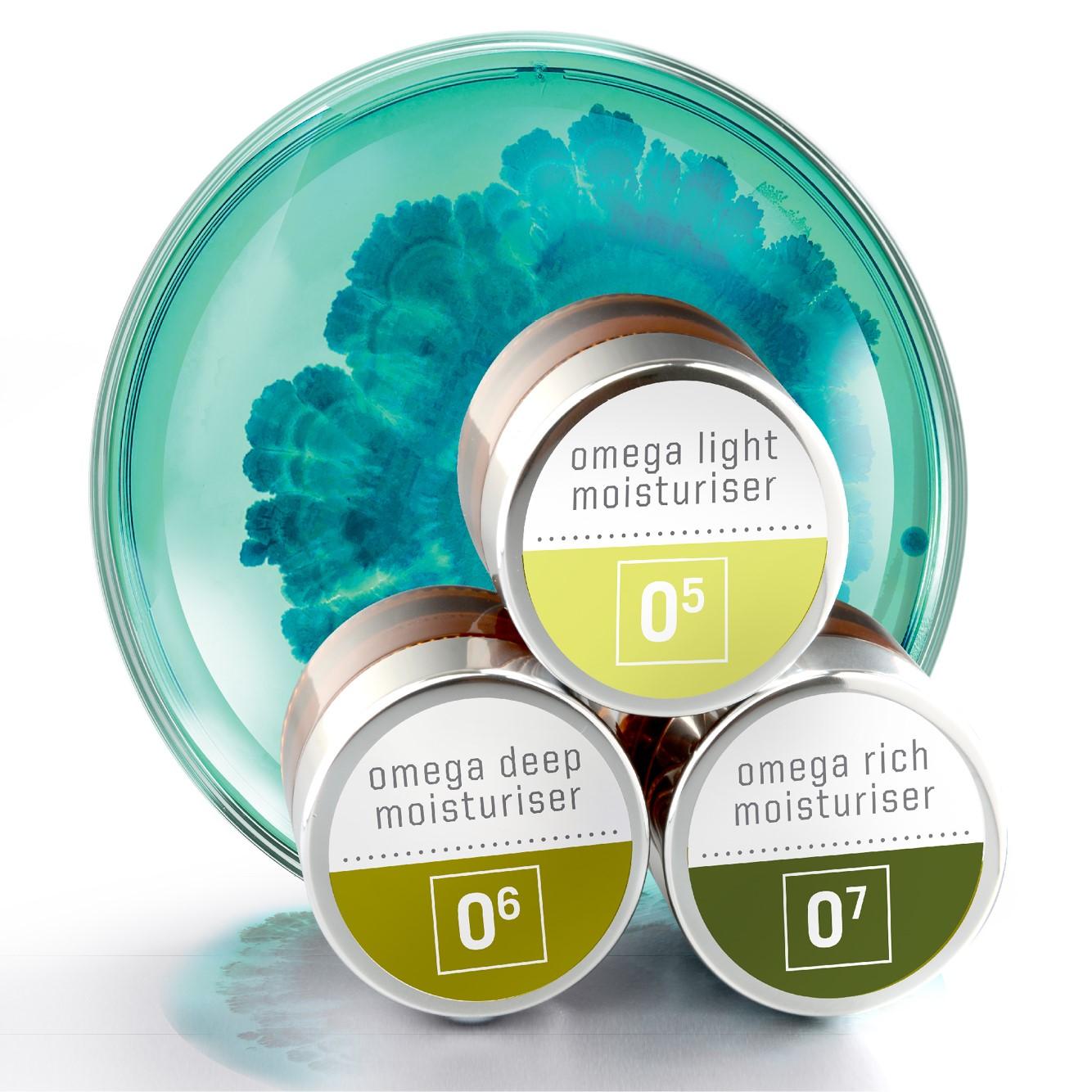 Three containers of moisturizer in front of a circular glass dish with a teal and green abstract pattern. Each container is labeled: "omega light moisturiser 05," "omega deep moisturiser 06," and "omega rich moisturiser 07.