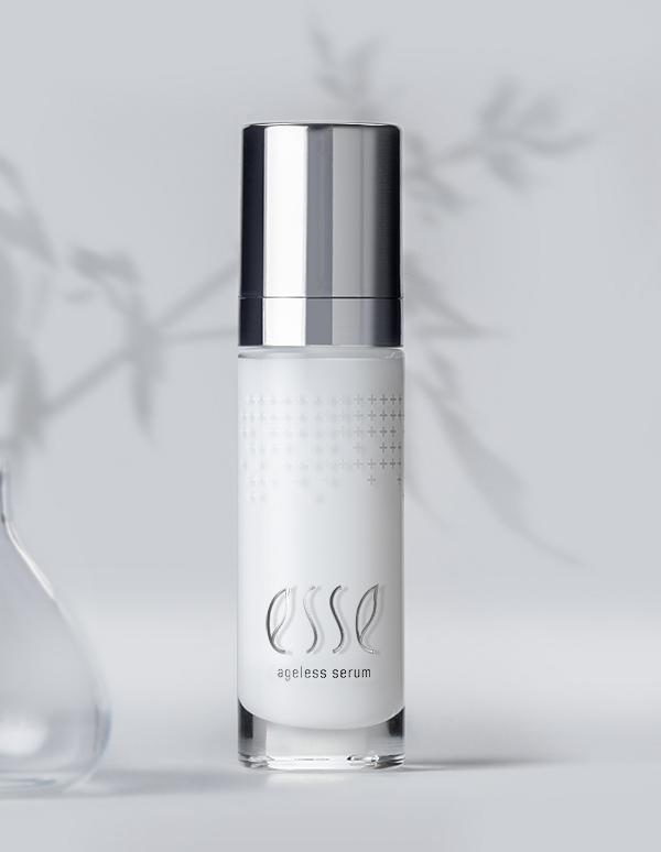 A sleek, white bottle of Esse Ageless Serum with a metallic silver cap is showcased against a soft, white backdrop adorned with blurred botanical patterns. The bottle prominently features the brand name "Esse" and the phrase "Ageless Serum" in a subtle, elegant font.