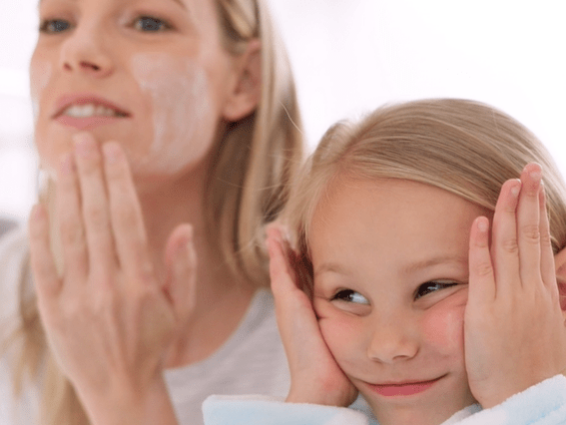 A woman is applying skincare cream to her face while smiling. Next to her, a young girl with long blonde hair is mimicking the action, touching her cheeks with a playful smile. Both are wearing casual, light-colored clothing, and the background is softly blurred.