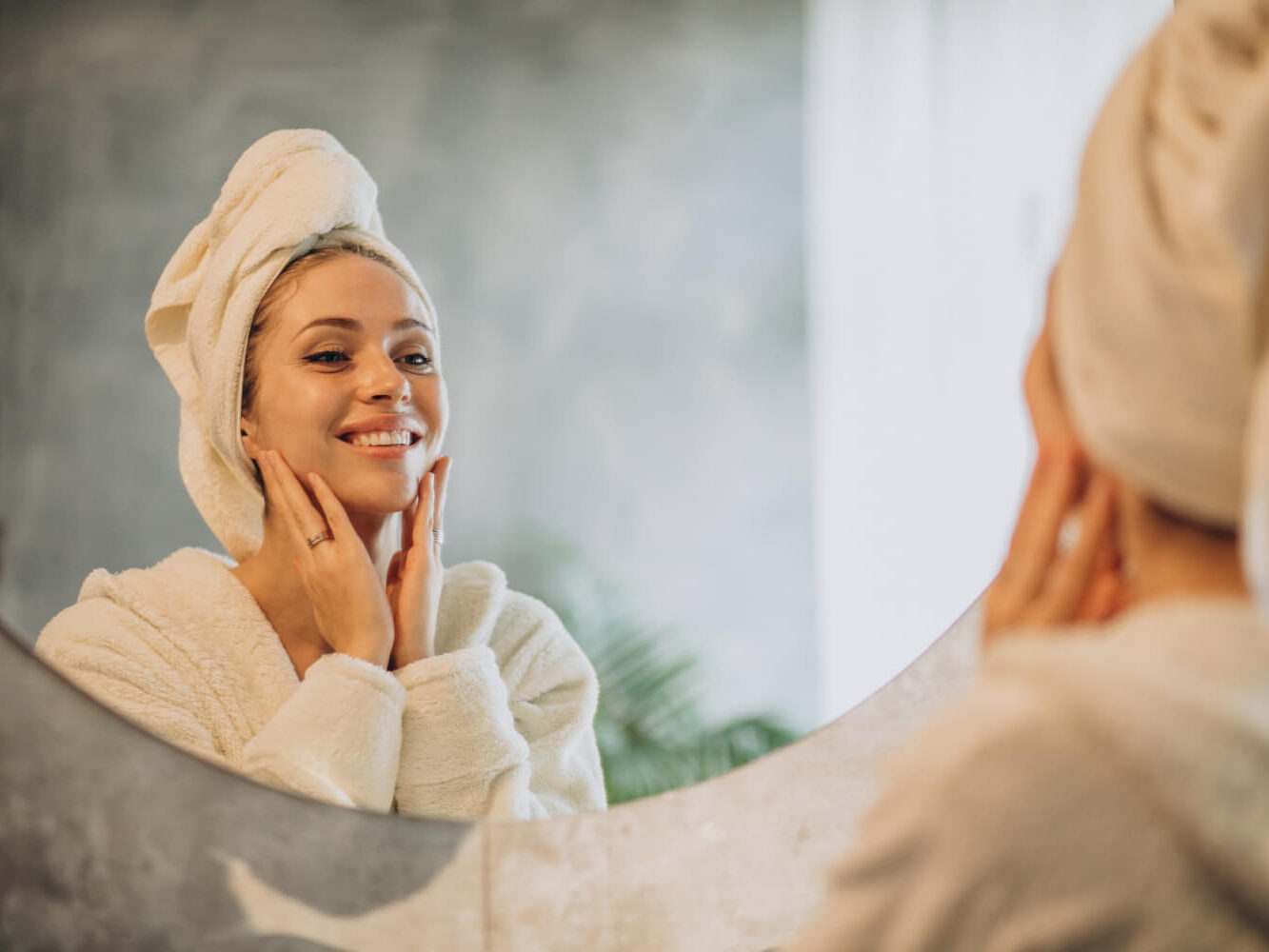 A person wrapped in a towel smiles at their reflection in a bathroom mirror while gently touching their face. The background features a leafy plant and soft, natural lighting.